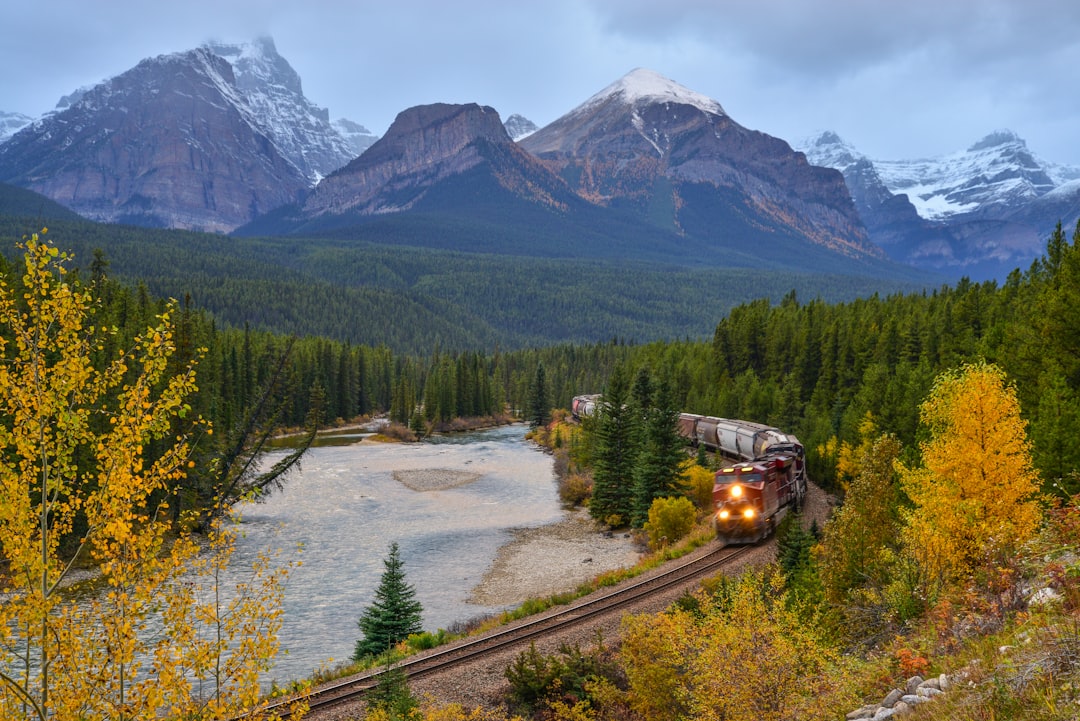 Train curving along a river valley beneath autumn trees and snowy peaks