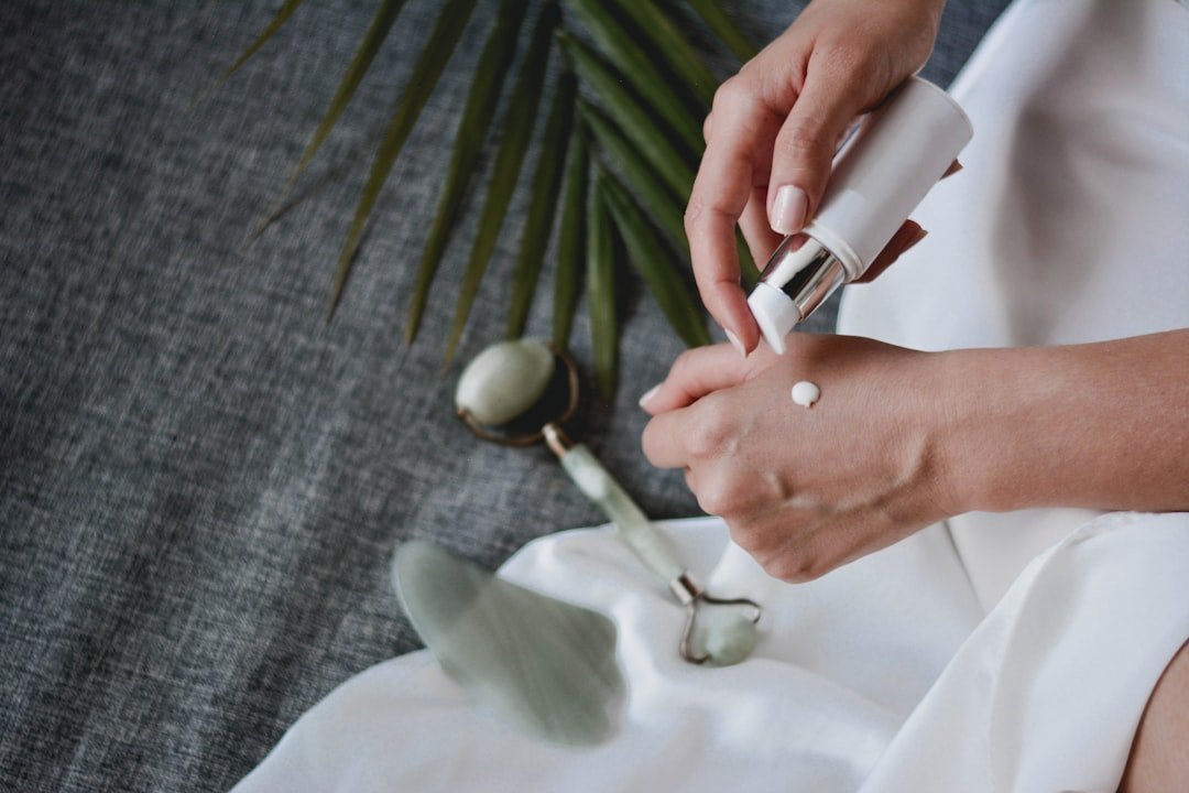 Hands dispensing facial moisturizer from a pump bottle with jade skincare tools nearby