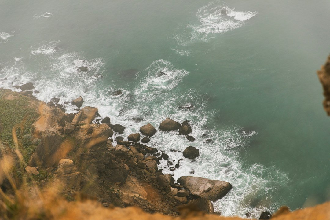 Turquoise waves breaking on boulders below rocky cliffs seen from above