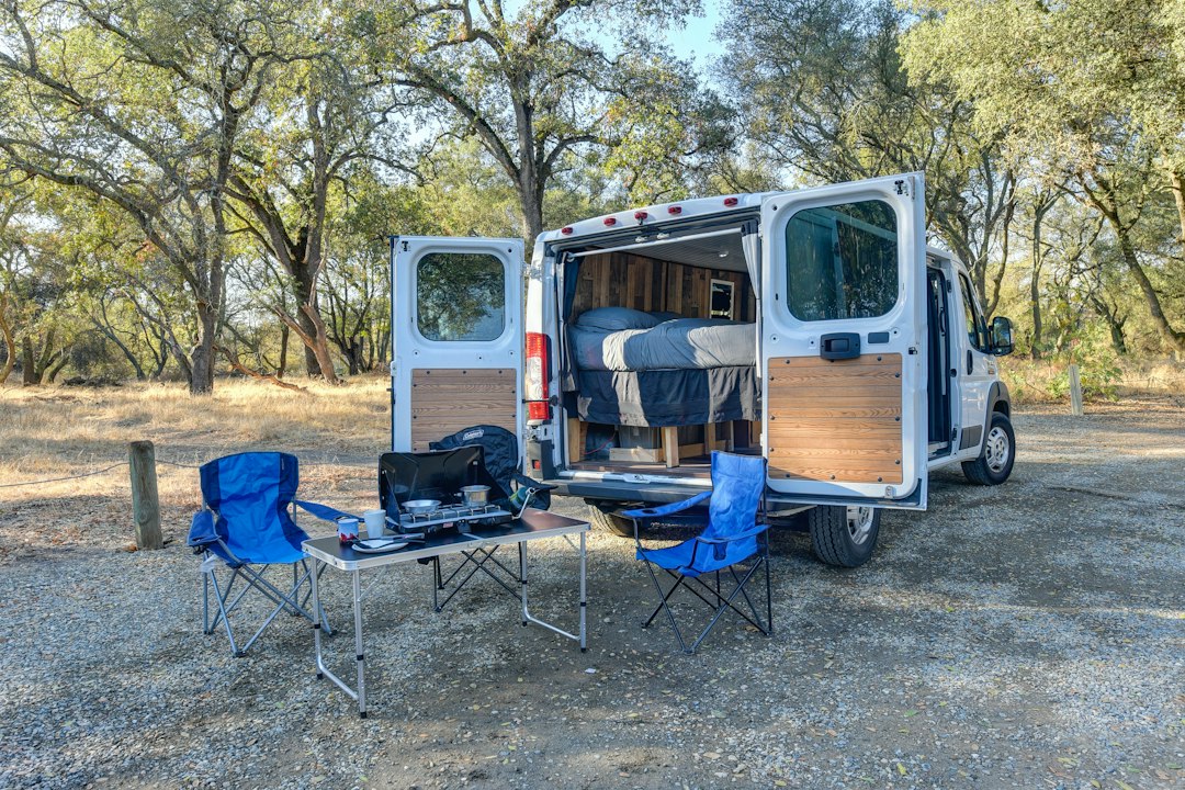 Camper van parked at a wooded campsite with camp chairs and a stove set up outside